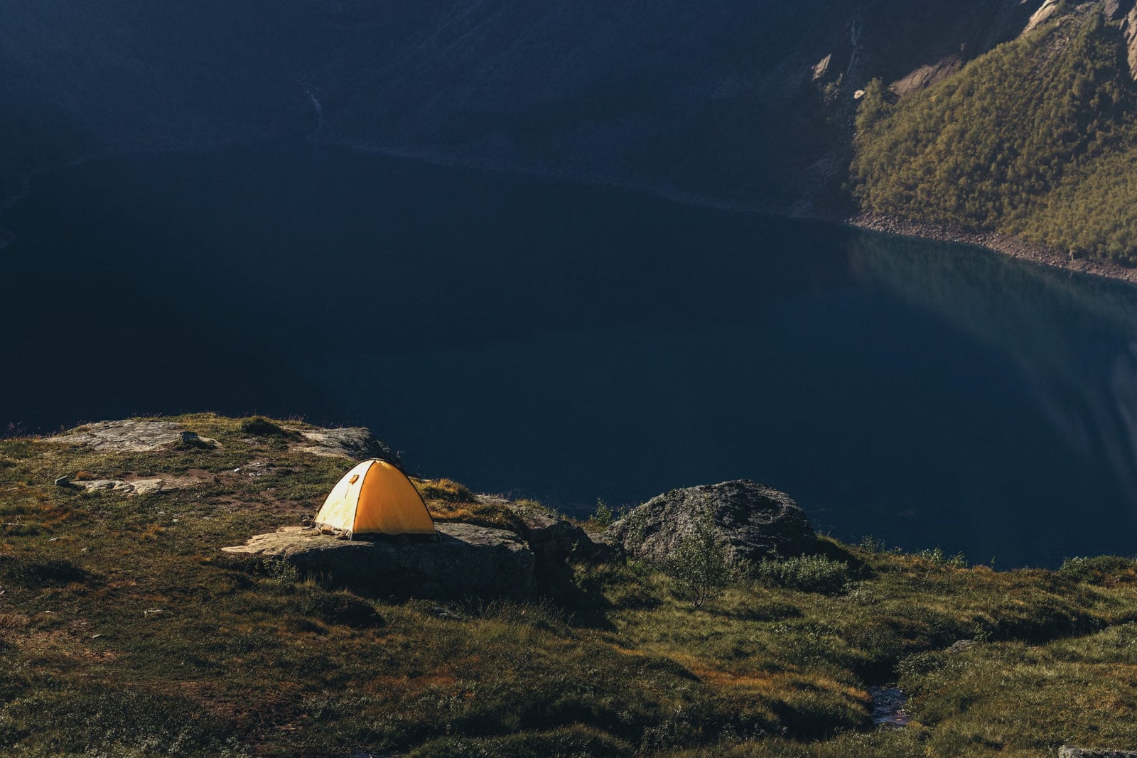 A yellow tent on a grassy ledge high above a dark, still fjord
