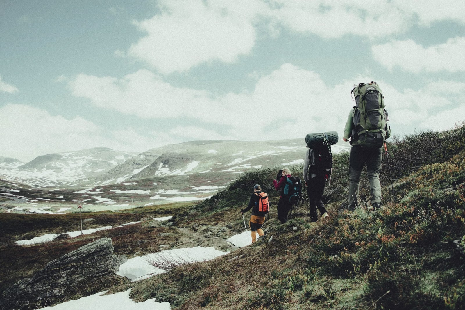 A small group with large backpacks traversing a green mountainside with patches of snow