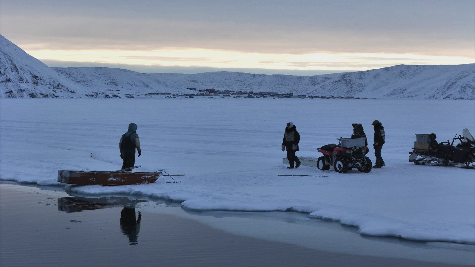 People crossing a frozen fjord at dusk, with snow-covered hills and a small settlement in the distance