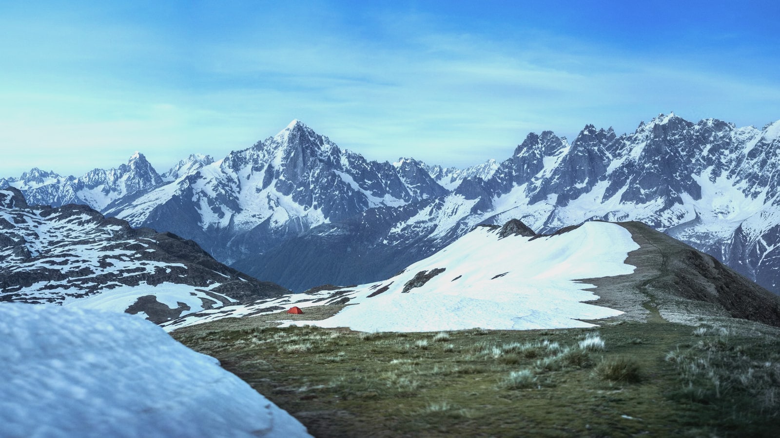 A lone red tent on a snowy ridge with a wide range of snow-covered peaks beyond